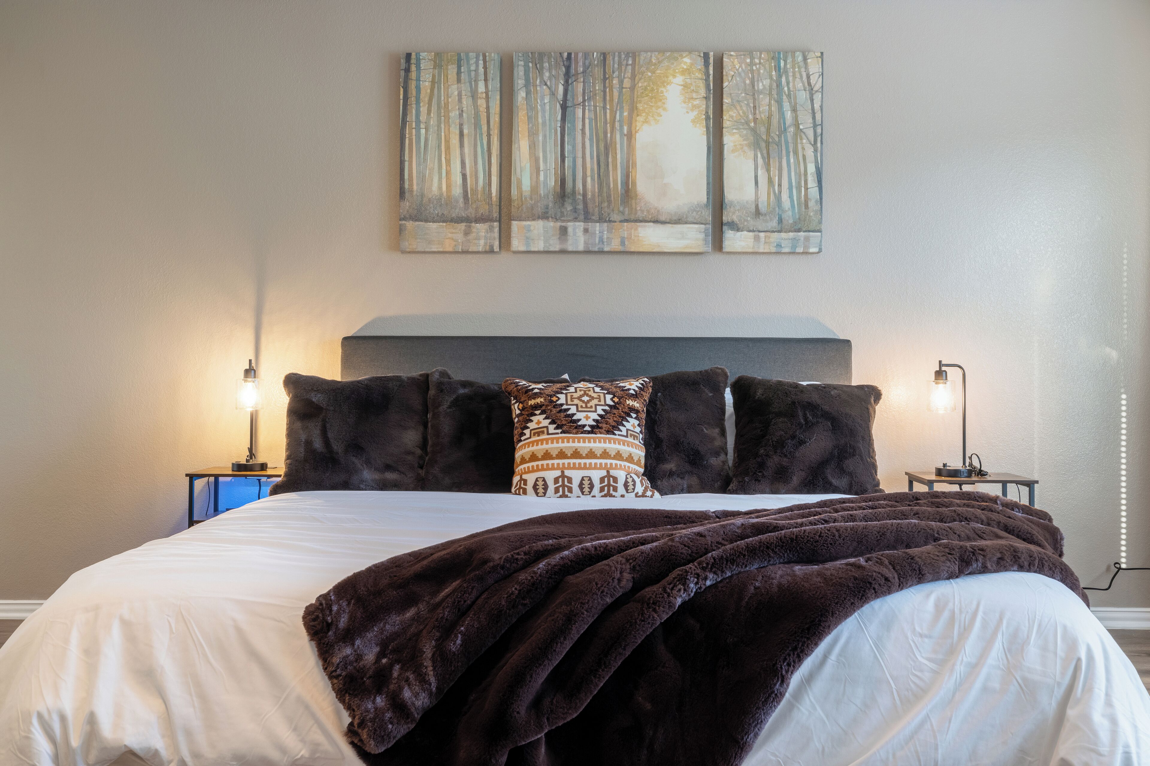Un lit bien fait, recouvert d’une couverture marron foncé, avec un oreiller à motifs et une tête de lit. Deux lampes de chevet sont placées de chaque côté du lit.