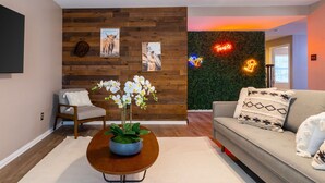 A modern living room with a wooden accent wall, a flat-screen TV, a sofa with patterned cushions, and a coffee table with a floral arrangement. - 5BR Retreat: Arcade, Cowboy Pool & Volleyball Oasis in Huge Backyard (Round Rock)