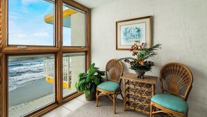 A room with a large window overlooking the beach, wicker chairs with cushions, a small table with a potted plant, and a framed picture on the wall.