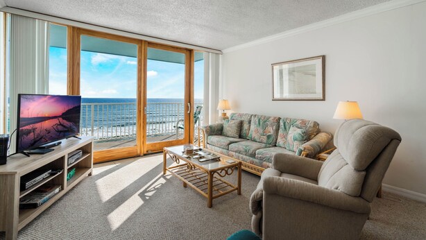 A living room with a television, sofa, and coffee table, a balcony with a view of the sea, and a framed picture on the wall.