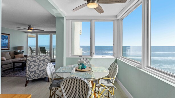 A dining area with a view of the ocean, featuring a round table, wicker chairs, and a ceiling fan.