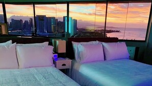 Habitación de hotel con dos camas, una mesita de noche y un amplio ventanal con vistas a la ciudad al atardecer.