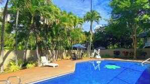 Una piscina con superficie de agua cristalina azul, rodeada de exuberante vegetación y un área sombreada con una silla.