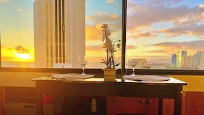 Una mesa de comedor con dos copas de vino y un jarrón de flores, frente a una vista al atardecer con edificios al fondo.