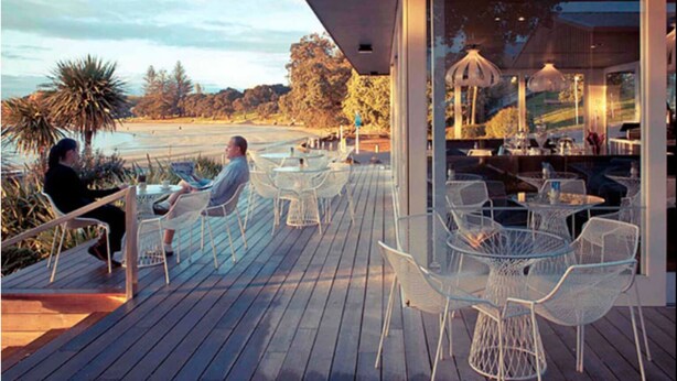 An outdoor dining area with glass tables and chairs, overlooking a beach and ocean.