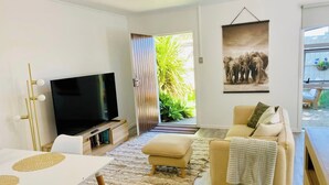 A modern living room with a flat-screen TV, a beige sofa, a dining table, and a large window with a view of greenery.
