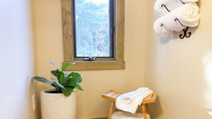 Ein Badezimmer mit einer Topfpflanze, einem kleinen hölzernen Hocker mit Handtüchern und einem Fenster mit Blick auf Bäume.