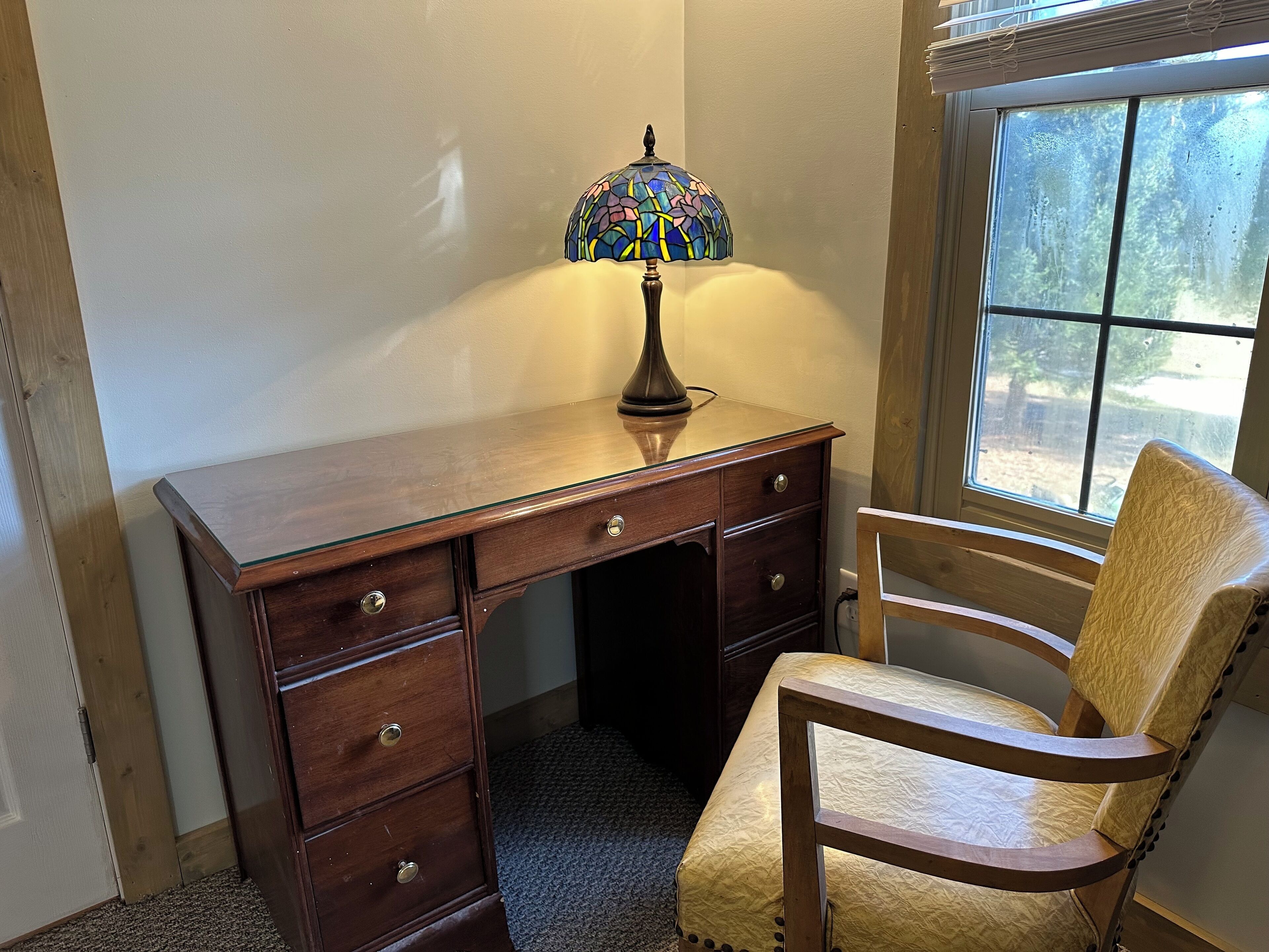 A corner desk with a stained glass lamp, a wooden chair, and a window with a view of trees.