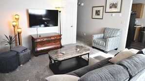 A living room with a flat-screen TV, a coffee table, a sofa, a patterned armchair, and a wooden dresser.