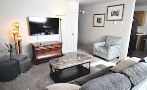 A living room with a flat-screen TV, a coffee table, a sofa, a patterned armchair, and a wooden dresser.