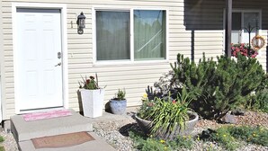 A house with a white door, two windows, a front porch, and a garden with various plants and flowers.