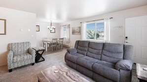 A living room with a gray sofa, a patterned armchair, a coffee table, and a dining area in the background.