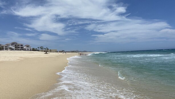 Ein Strand mit blauem Himmel, sandigem Ufer und Gebäuden im Hintergrund.