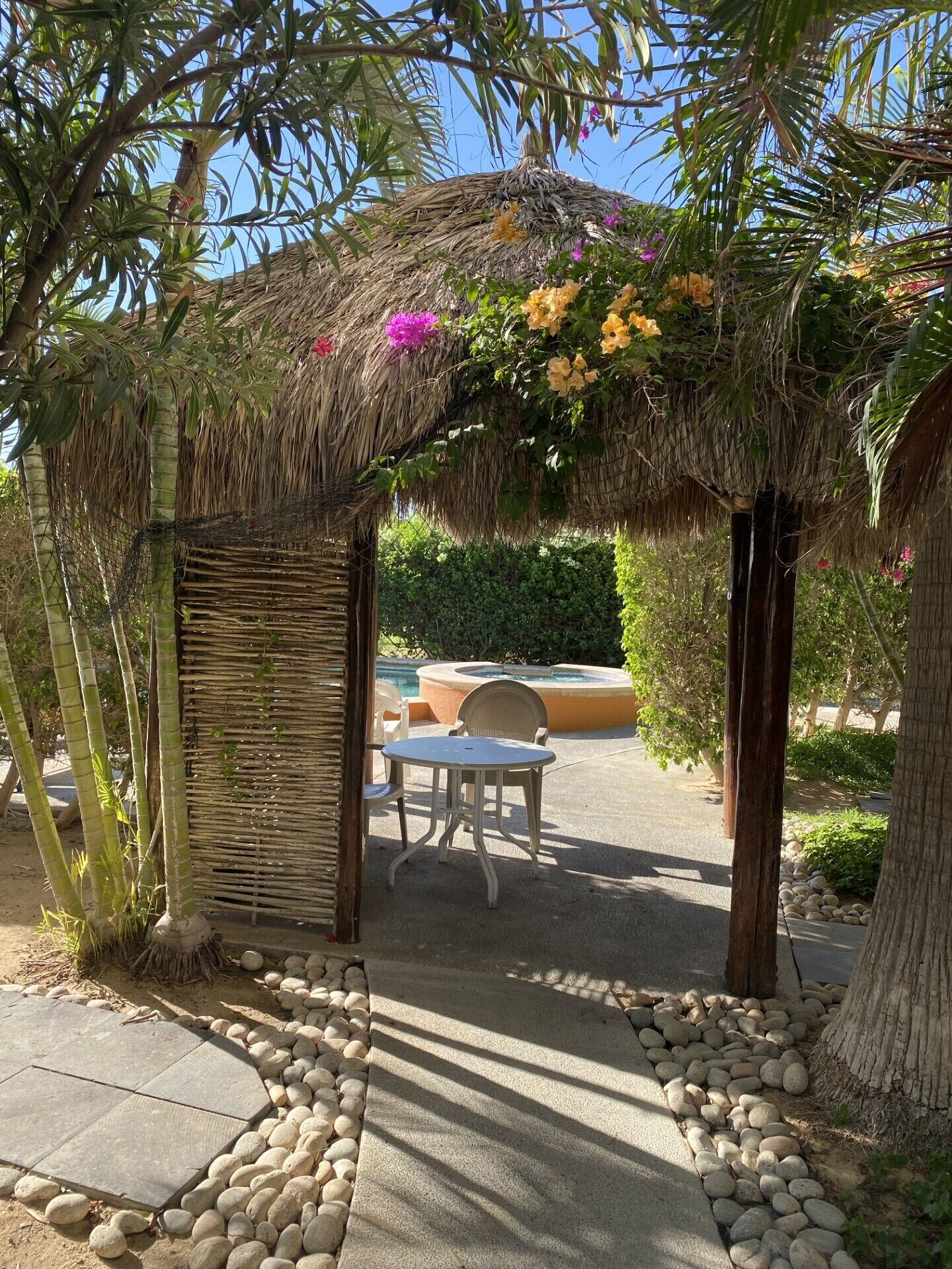 A thatched gazebo with a table and chairs underneath, surrounded by a stone pathway and greenery.