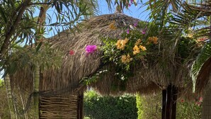 Ein strohgedecktes Gartenhaus mit Tisch und Stühlen, umgeben von einem Kiesweg und Grünpflanzen.