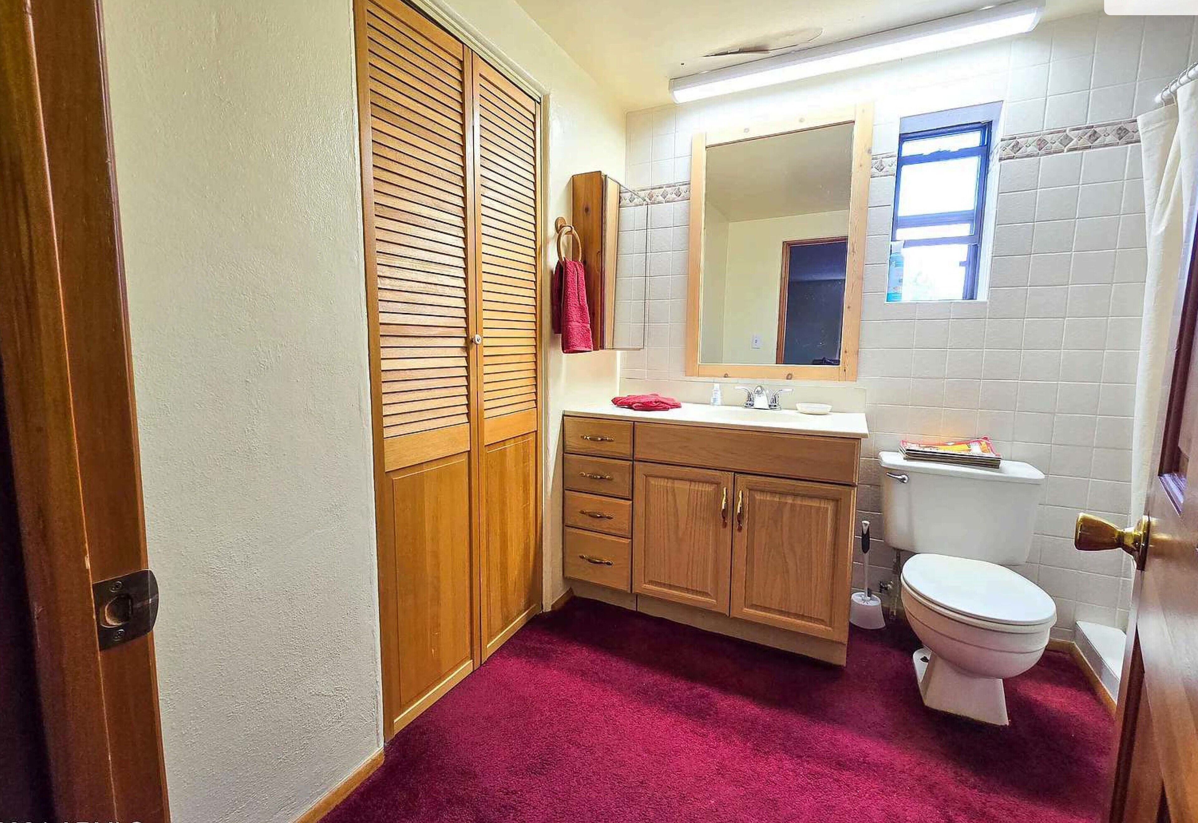 Une salle de bain avec des meubles en bois, des toilettes blanches, un miroir et un tapis rouge.