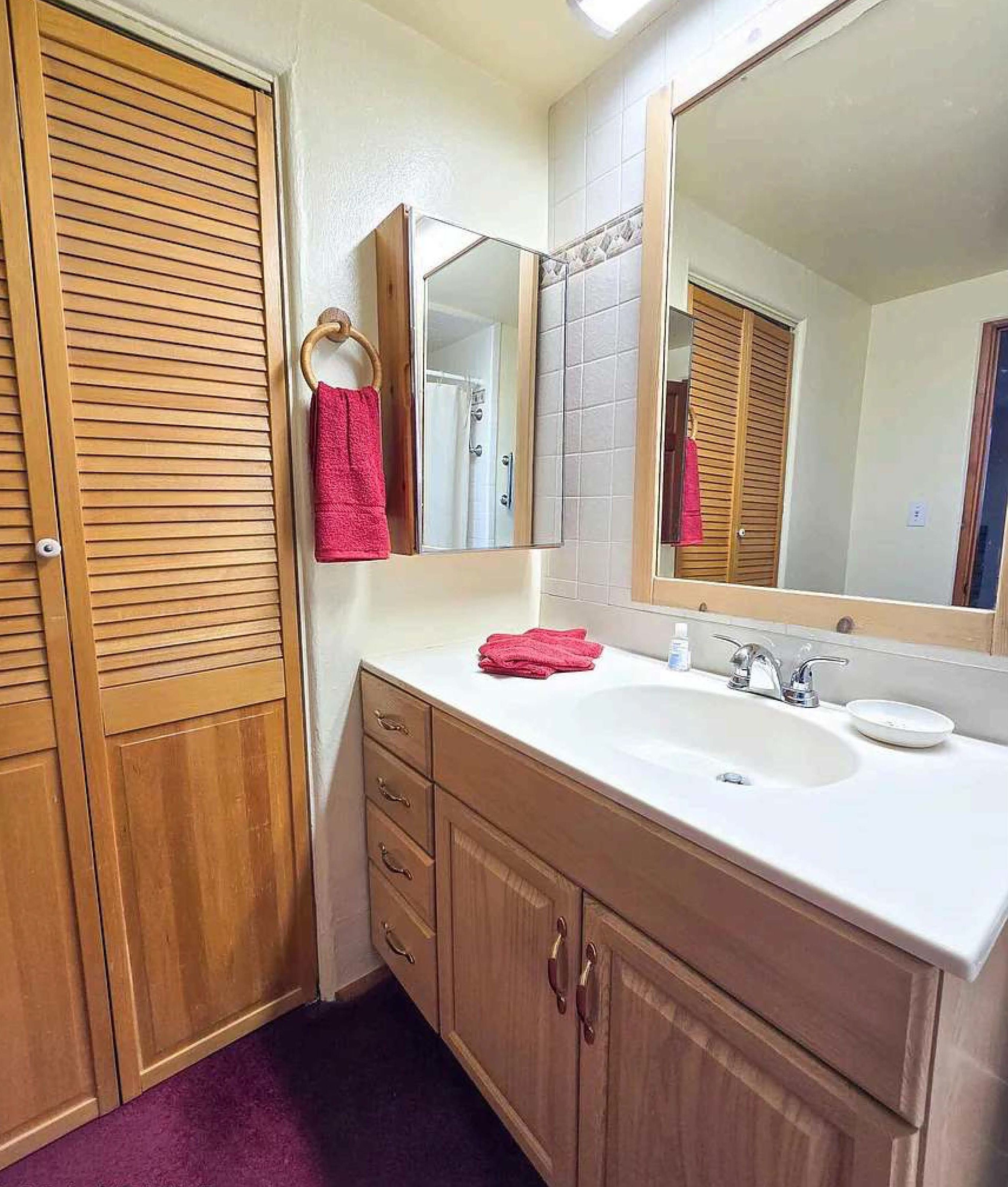 Un baño con armarios de madera, un lavabo blanco, un espejo y una toalla roja colgada en un perchero.