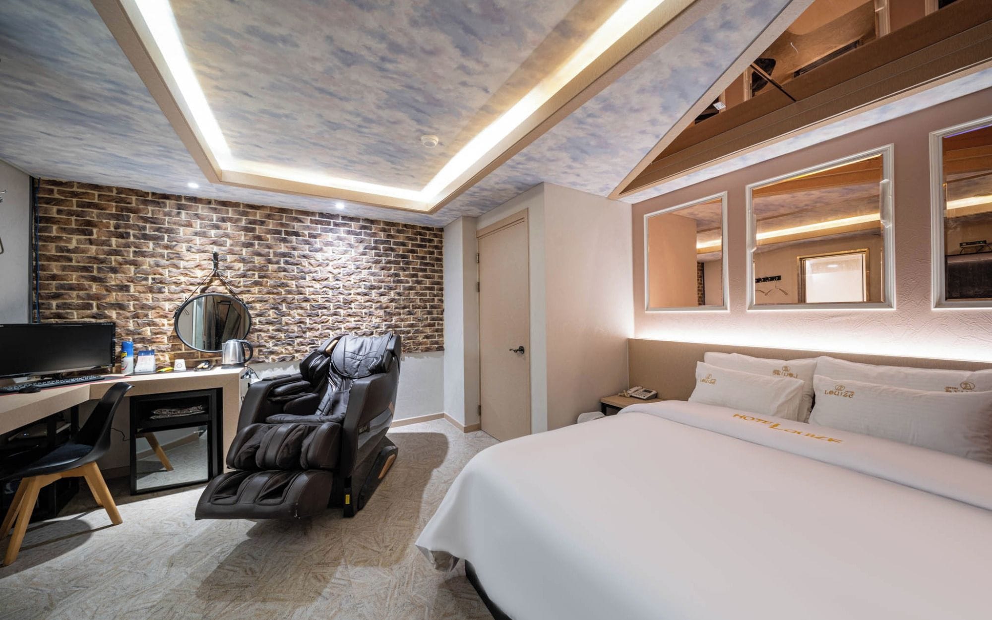 Quarto de hotel com parede de tijolos, poltrona de massagem, escrivaninha com computador e cama com roupa de cama branca.