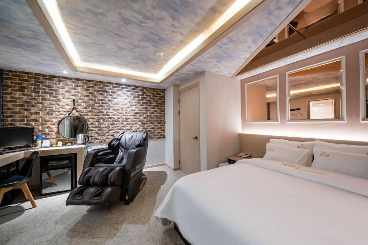A hotel room with a brick wall, a massage chair, a desk with a computer, and a bed with white bedding.