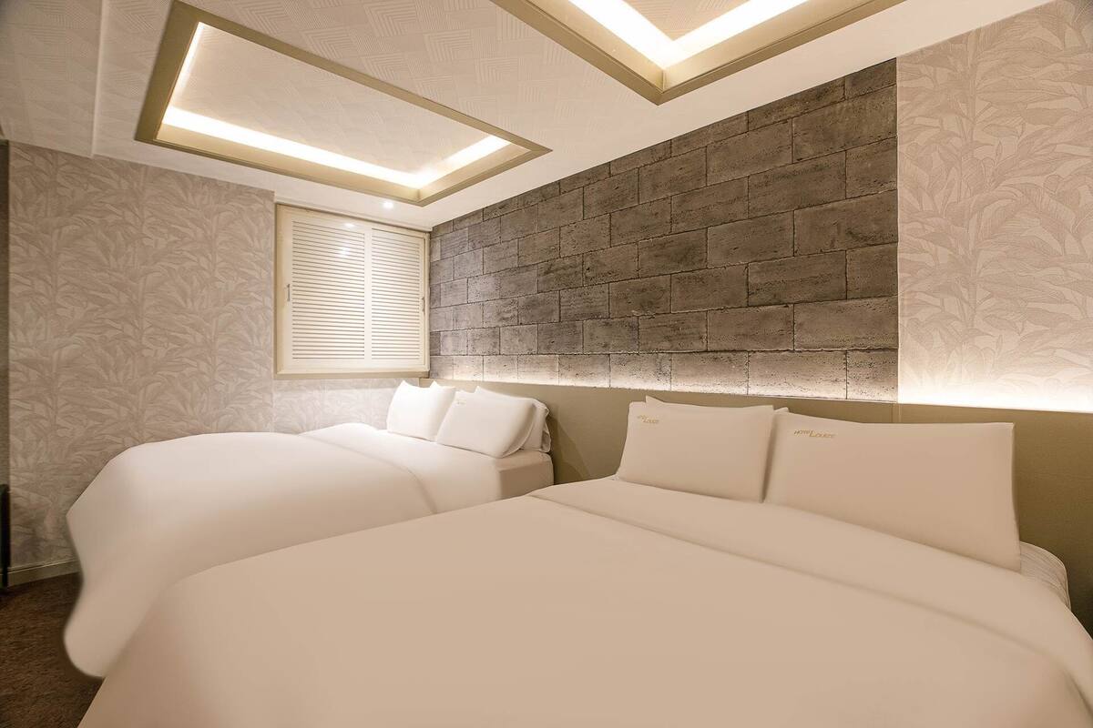 A bedroom with two beds, a sloped ceiling with integrated lighting, a brick accent wall, and a window with blinds.