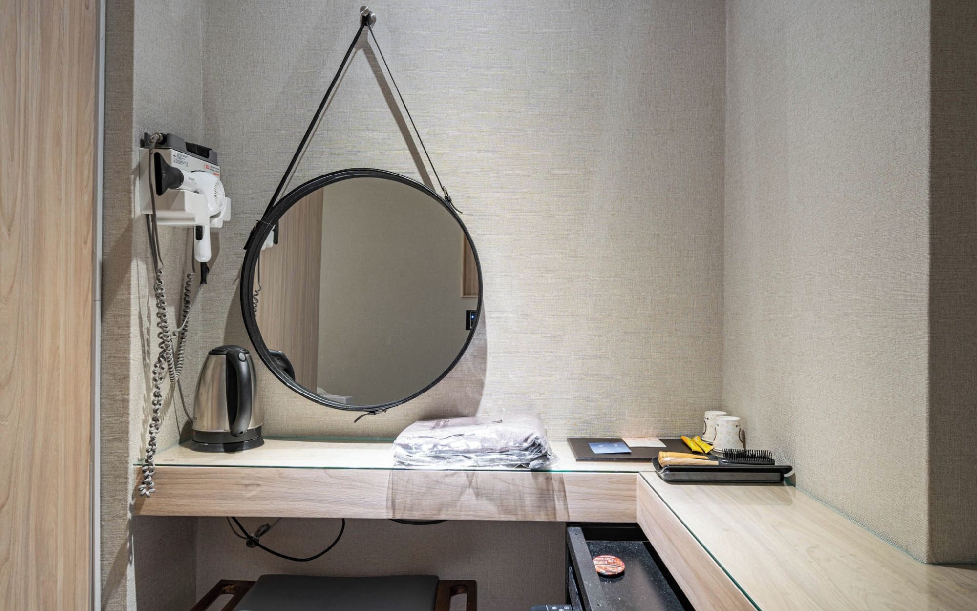 A compact hotel room with a small desk, a round mirror, a kettle, and a portable grill.