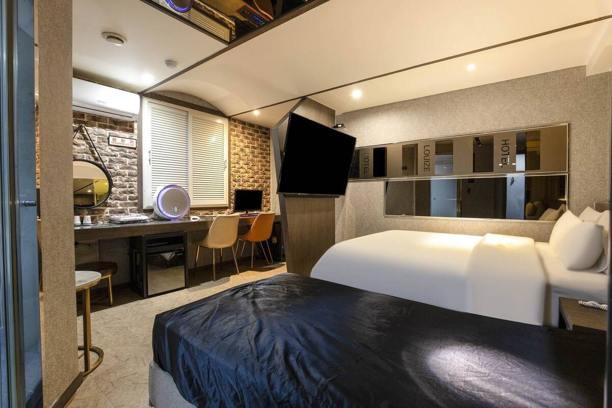 A modern hotel room with a large bed, a desk with a computer, a flat-screen TV, a mirror, and a stone wall.