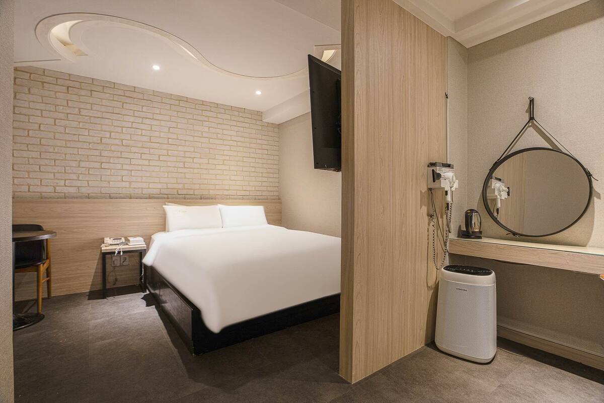 A modern hotel room with a brick-patterned headboard, a large bed, a flat-screen TV mounted on the wall, a round mirror, and a Samsung air purifier.
