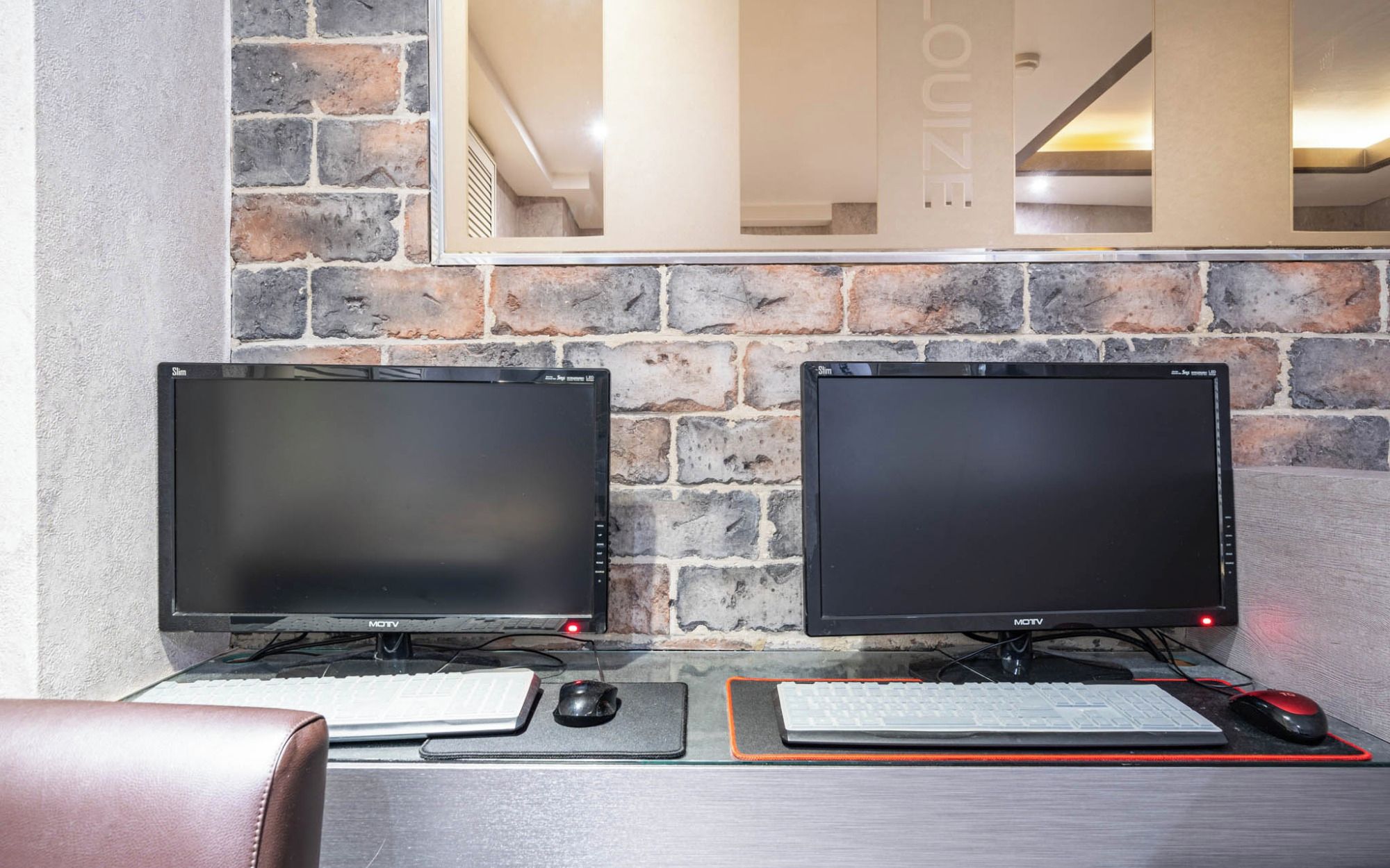 Two monitors on a desk with a keyboard and mouse, set against a stone wall with a mirror reflecting the room.
