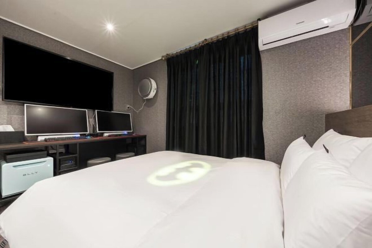A hotel room with a bed, a TV, a desk with a computer, and an air conditioning unit.