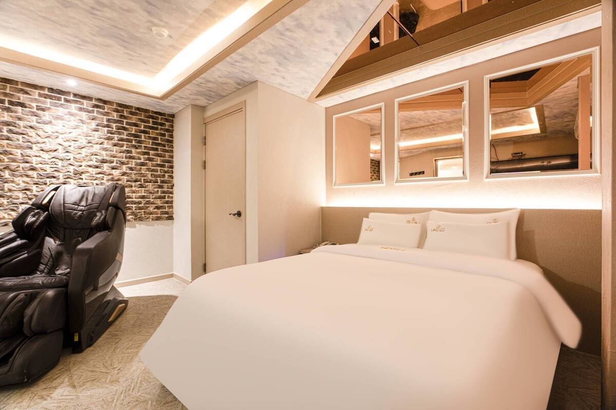 A hotel room with a massage chair, a bed with white linens, a brick wall, and a wooden ceiling.