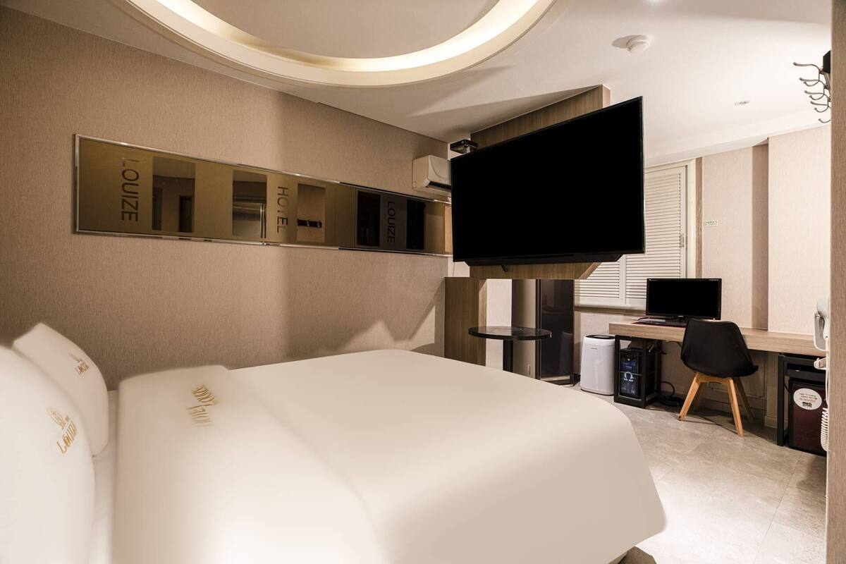 A hotel room with a large bed, a desk with a computer, a flat-screen TV, and a minibar.