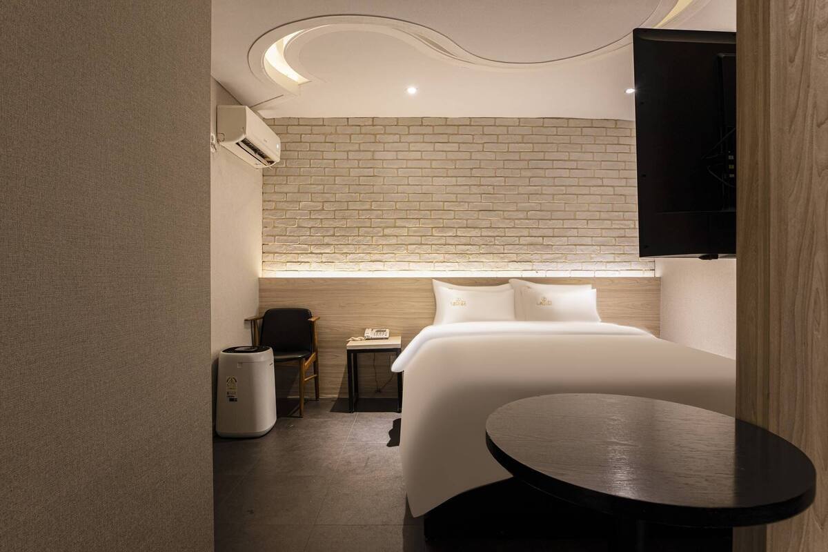A compact hotel room with a brick-patterned wall, a bed, a small table, a chair, and a TV mounted on the wall.