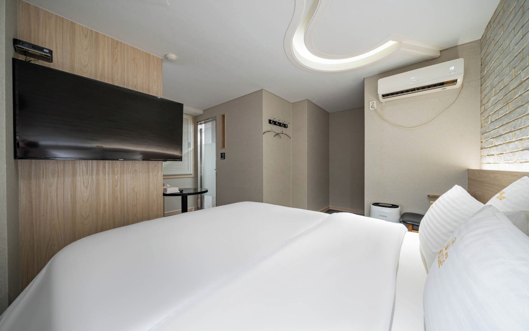 Ein Hotelzimmer mit Bett, einem Flachbildfernseher an der Wand, einer Wandklimaanlage und einem kleinen Tisch mit Stuhl.