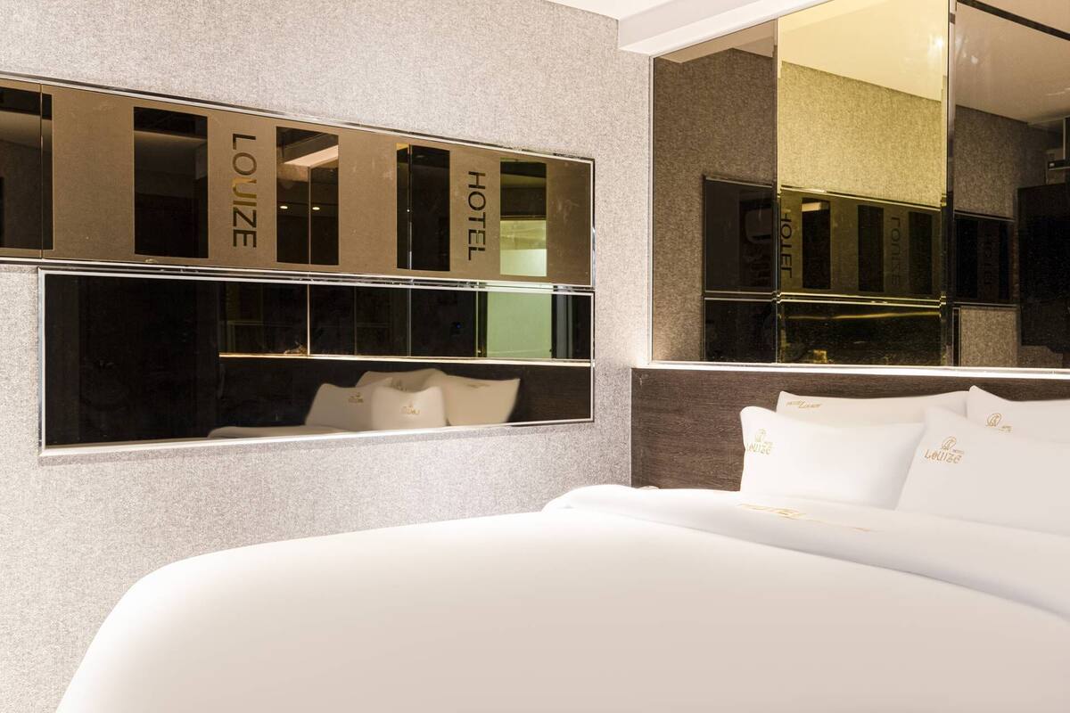 A modern hotel room with a large bed, a mirror reflecting the room, and the hotel's name on the mirror.