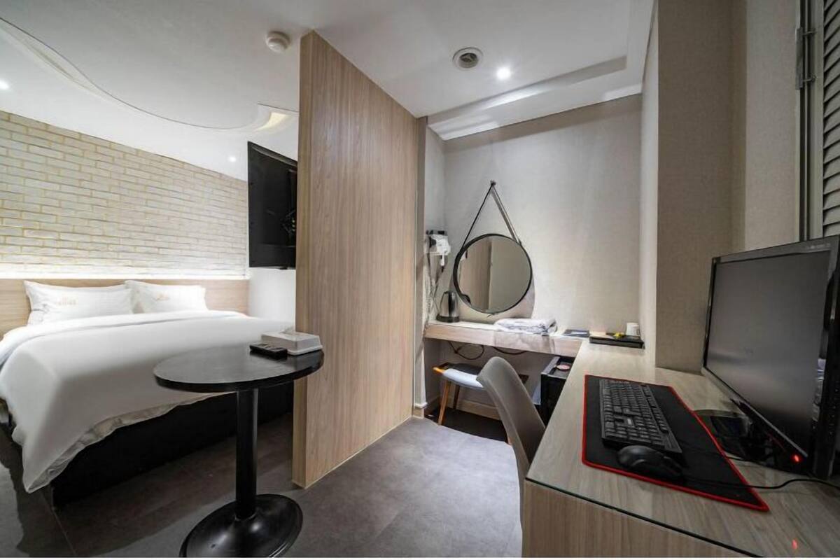 A modern hotel room with a bed, a desk with a computer, a chair, and a small table.