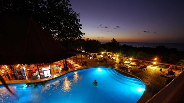 A nighttime swimming pool with a curved edge, surrounded by lounge chairs and a covered area with tables and chairs.