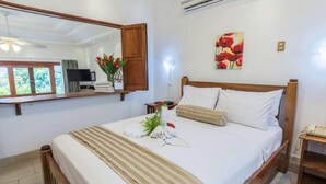 A neatly made bed with white linens and a striped blanket, a wooden headboard, a bedside table with a lamp, a mirror, and a painting of red flowers on the wall.