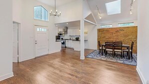 A spacious open-plan living area with a dining table and chairs, a kitchen in the background, and a brick accent wall.