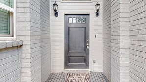A gray door with a welcome mat, two wall-mounted lanterns, and a window above.