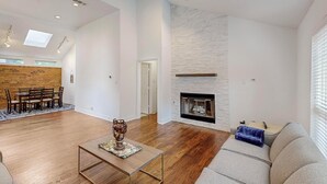 A spacious living room with hardwood floors, a fireplace, a dining area with a brick accent wall, and a large window providing natural light.