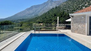 A swimming pool with a mountain view, a glass railing, and a stone building.