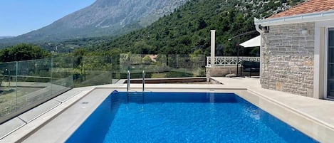 A swimming pool with a mountain view, a glass railing, and a stone building.
