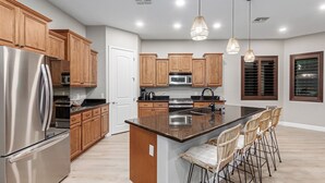 A modern kitchen with wooden cabinets, stainless steel appliances, a central island with bar stools, pendant lighting, and a window with blinds. - Modern South Mountain Heated Pool Trails Fire Pit (Phoenix)