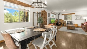 A spacious dining area with a long table, white chairs, and a chandelier. There is a fireplace with a stone mantle and a television on the wall. - Delightful 3-bedroom house in charming Flagstaff w/King Size Bed & Gas Log Stove (Flagstaff)