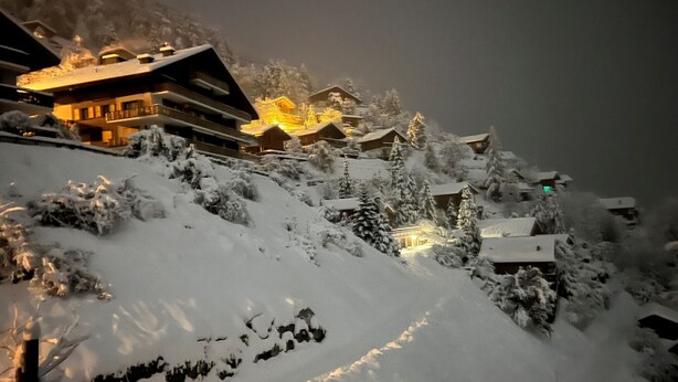 Una località montana innevata con diversi edifici, tra cui una struttura principale con balconi e un ingresso illuminato.