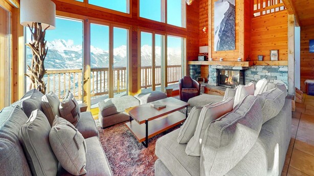 L’intérieur chaleureux d’un chalet en bois, avec une cheminée, de grandes fenêtres offrant une vue sur les montagnes et un coin salon confortable.