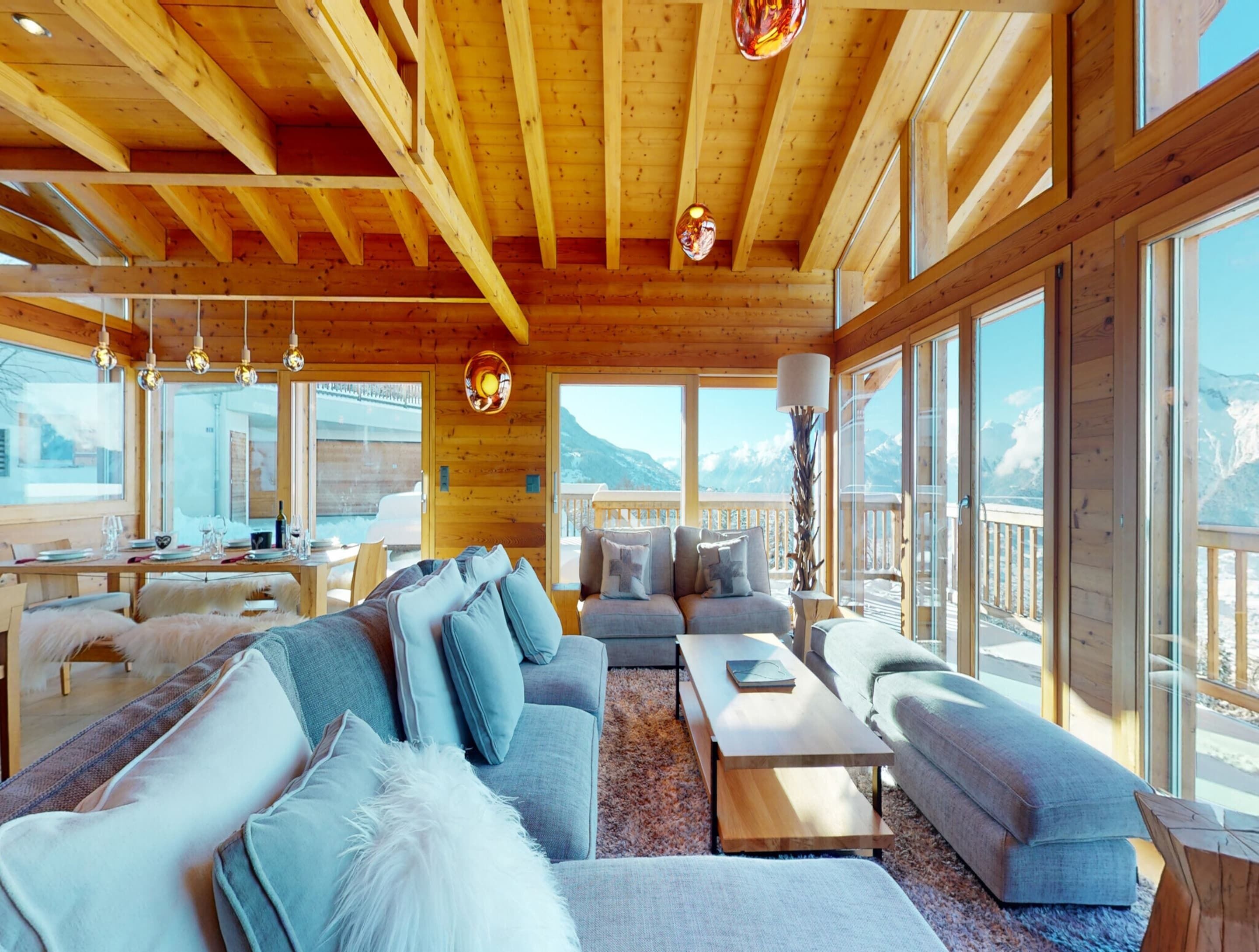 A spacious wooden interior with a sofa, coffee table, and dining area, featuring large windows and a view of mountains.