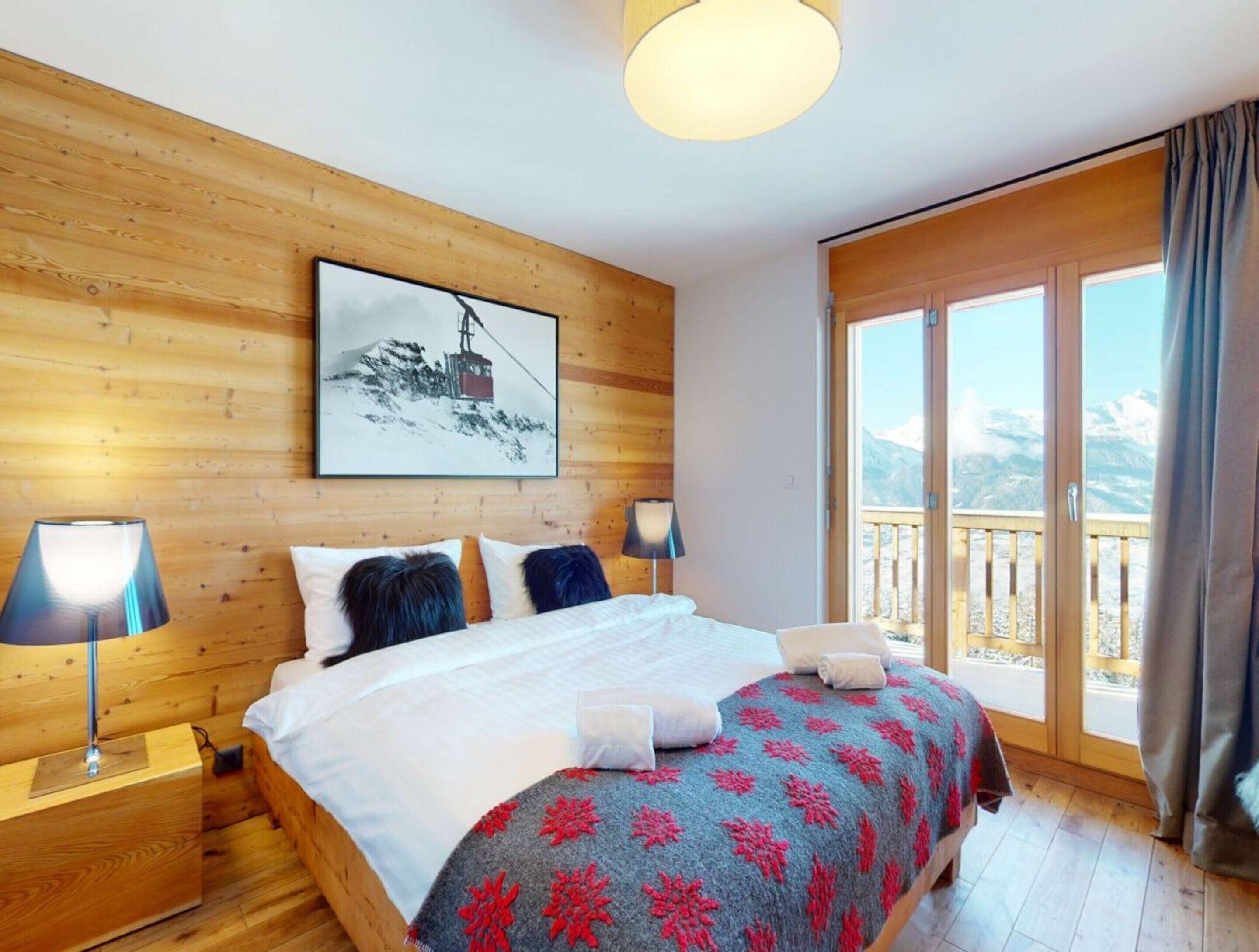 Una camera da letto con testiera in legno, un letto grande con coperta rossa e blu, due lampade ai comodini e una finestra vista montagna.