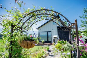 A metal archway with hanging planters, a house with a dark exterior and large windows, and a clear blue sky. - SONNET HIGASHIKAWA (Higashikawa)