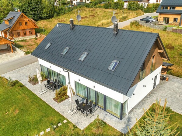 A modern two-story house with a sloped roof, large windows, and a patio area with outdoor furniture. - Comfortable Villa With Beautiful View (Cerny Dul)